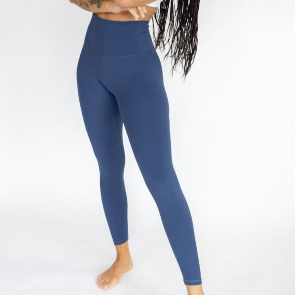 A person with long braided hair and tattoos stands barefoot, arms crossed, modeling DYM Athletics’ Ultra High Waist Flex & Flow Legging | SALE with a matching cropped top against a white background. Their head is out of frame.