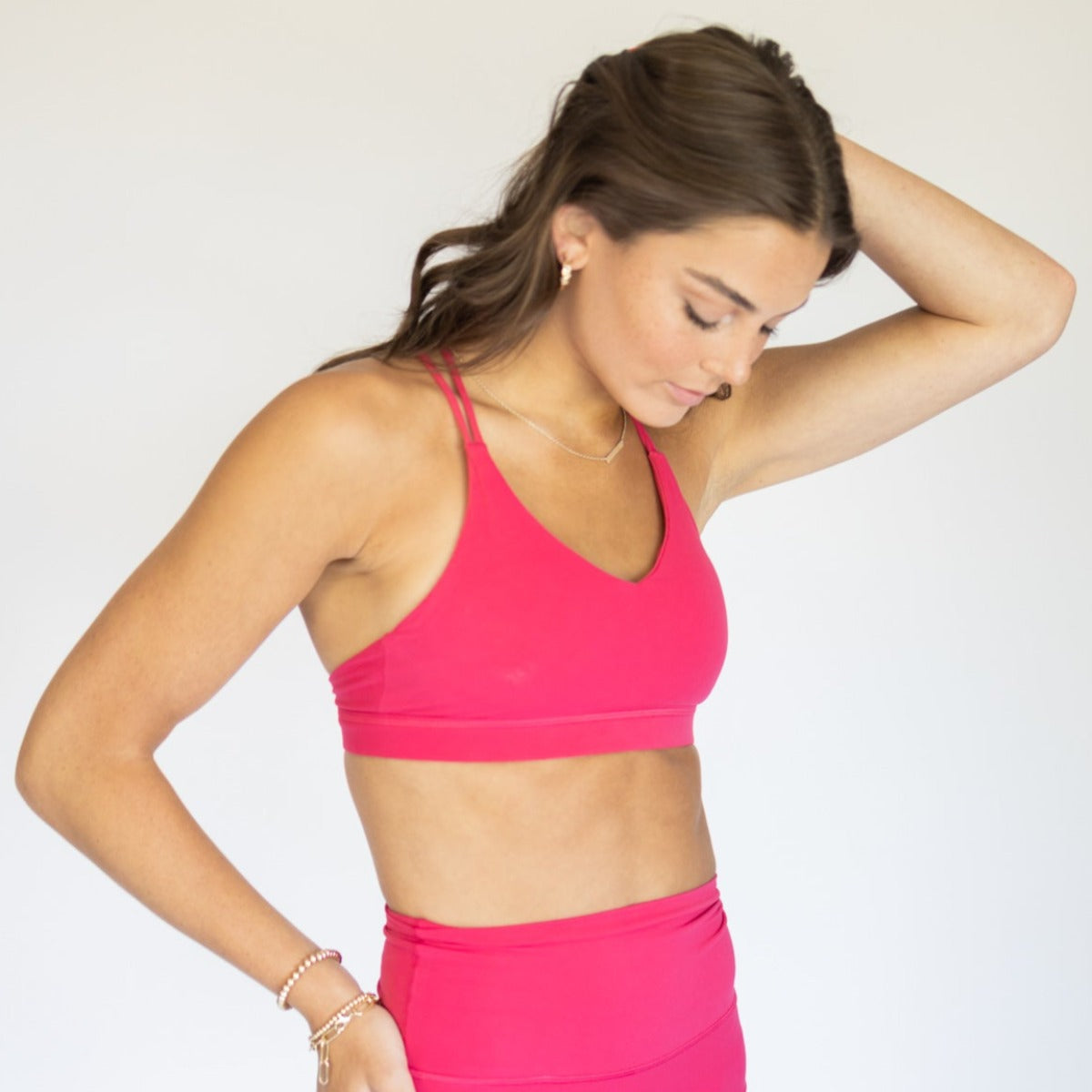 A woman with long brown hair models the DYM Athletics AIRE Ultra-Soft Cross Back Sports Bra (A-D Cup, Medium Support) in bright pink, standing against a plain light grey background with one hand behind her head.