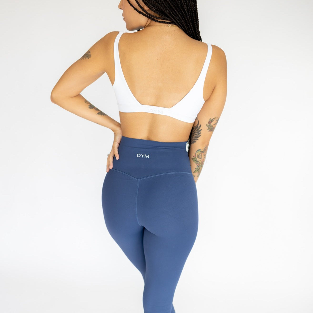 A woman with braided hair and arm tattoos is seen from behind, wearing a white sports bra and DYM Athletics Ultra High Waist Flex & Flow Leggings. She stands against a plain white background, one arm bent as she touches her hair.