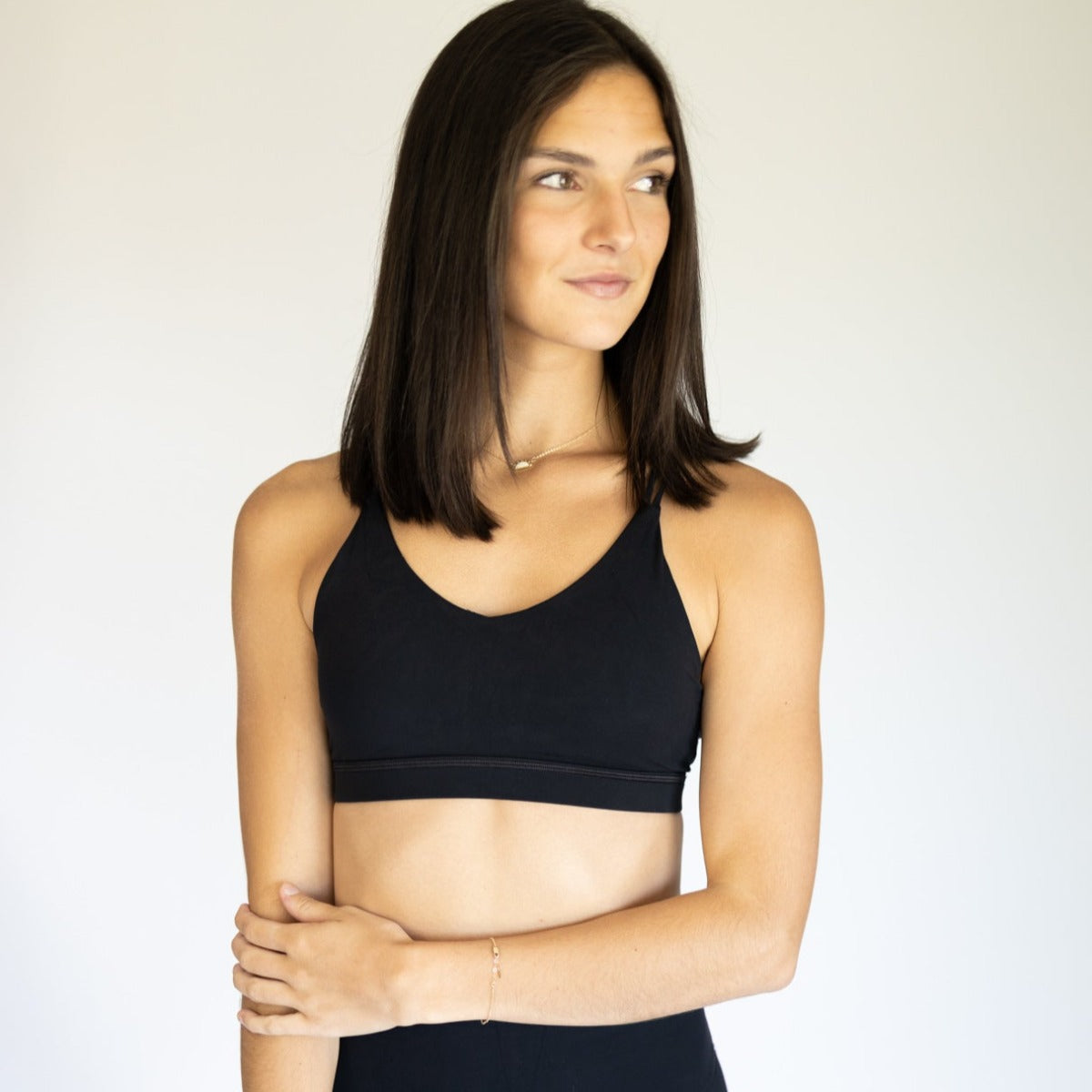 A woman with straight, shoulder-length brown hair wears the DYM Athletics AIRE Ultra-Soft Cross Back Sports Bra (A-D Cup, Medium Support) in black, styled with black pants. She stands against a light background, gazing right with a slight smile.
