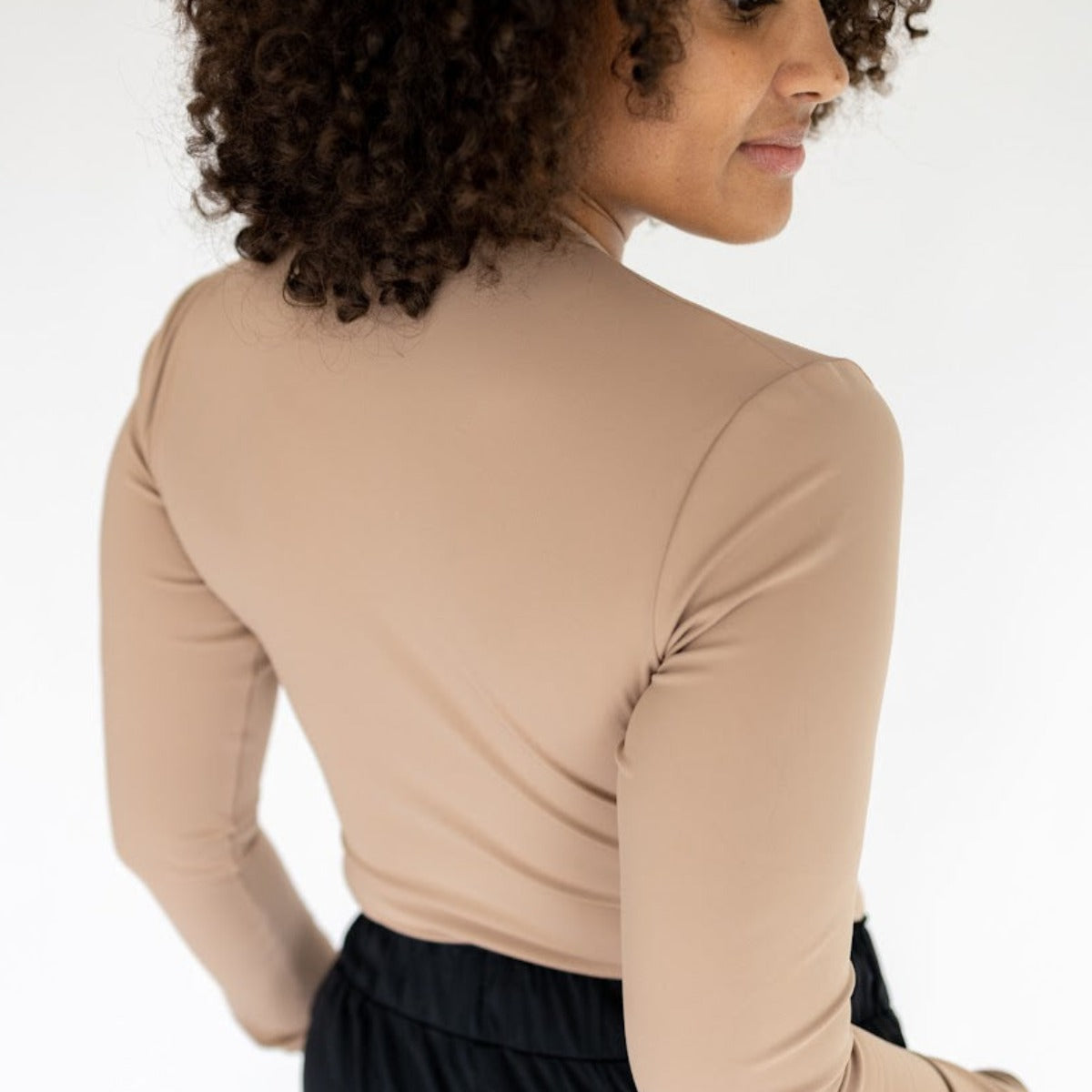 A woman with curly brown hair, viewed from behind, models the Lavish Shaping Bodysuit by DYM Athletics—featuring a compressive fit, full-coverage neckline, and black bottoms—against a minimalist white background.