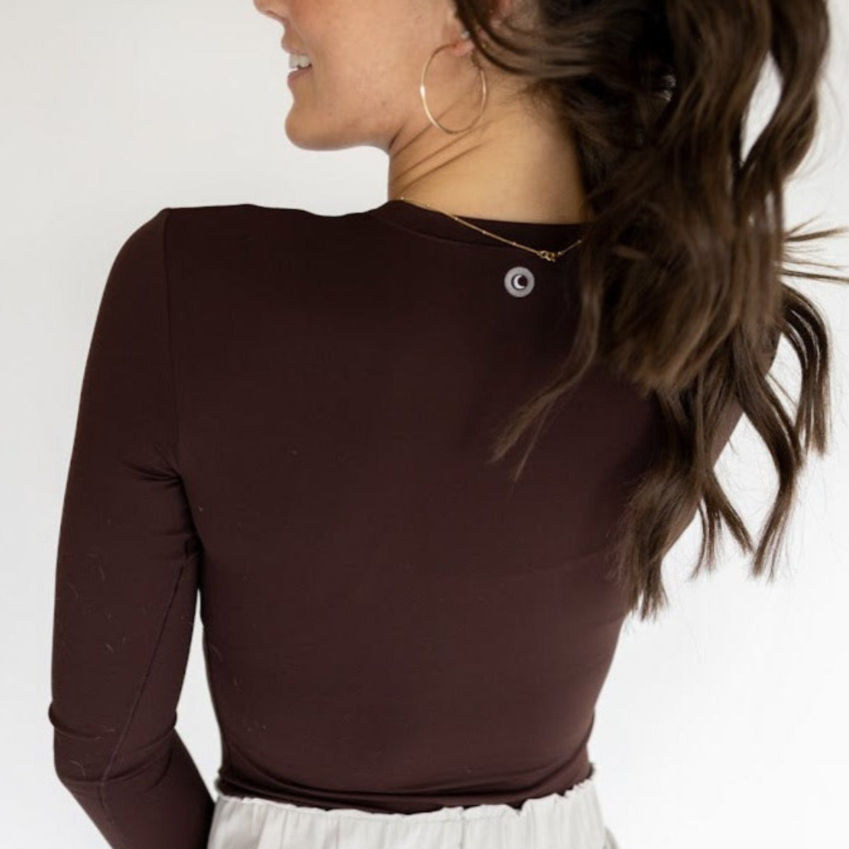 A woman with long, wavy brown hair in a ponytail is seen from behind, wearing the DYM Athletics Lavish Shaping Bodysuit in dark brown with a full coverage neckline and second-skin fit, paired with a light skirt and gold accessories against a white background.