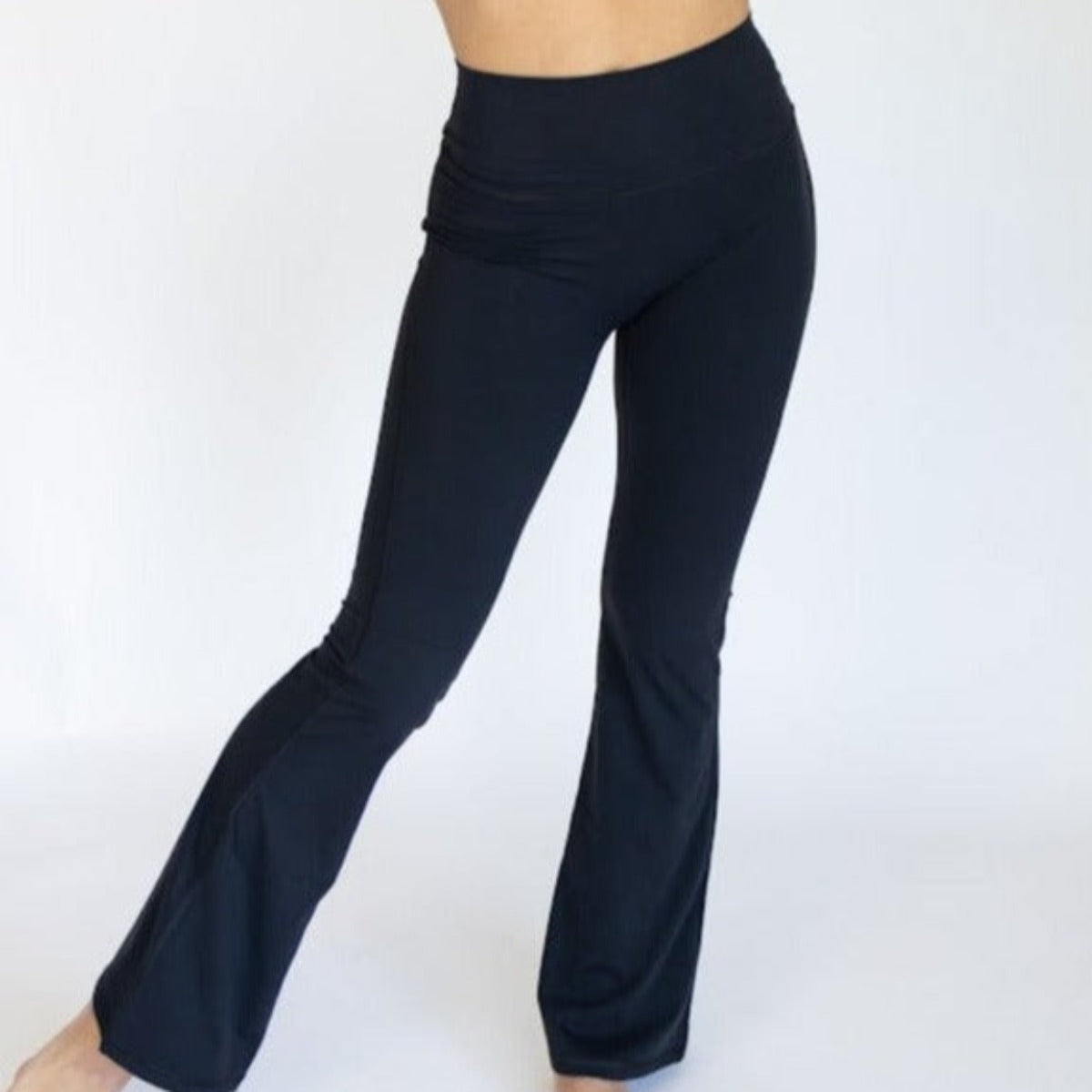 Wearing DYM Athletics' AIRE Ultra-Soft Fabulous Flare High Waist Pant | SALE in black, a person stands on one foot with the other leg slightly bent, toes touching the plain white floor. Only the lower body is shown; face and upper body are not visible.