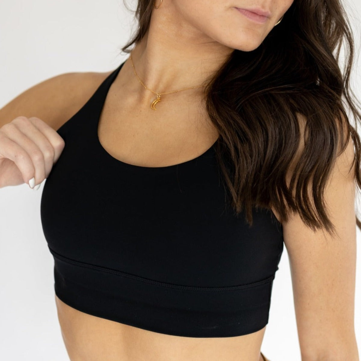 A woman with long brown hair models the DYM Athletics Zone Bra | A-D+ cup | High Support in black, paired with a gold crescent moon necklace. She poses sideways with one arm bent against a plain white background.