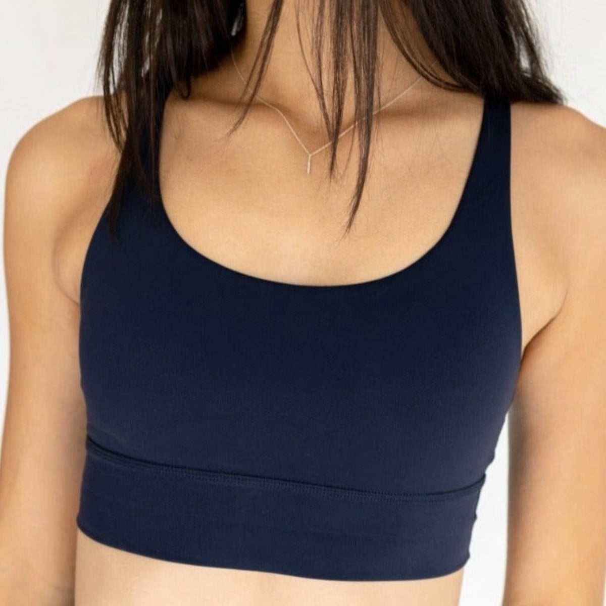 A woman with long brown hair wears the DYM Athletics Zone Bra | A-D+ cup | High Support in navy blue, paired with a delicate silver necklace. Made from moisture-wicking fabric. Her face isn’t visible; the background is plain and light-colored.