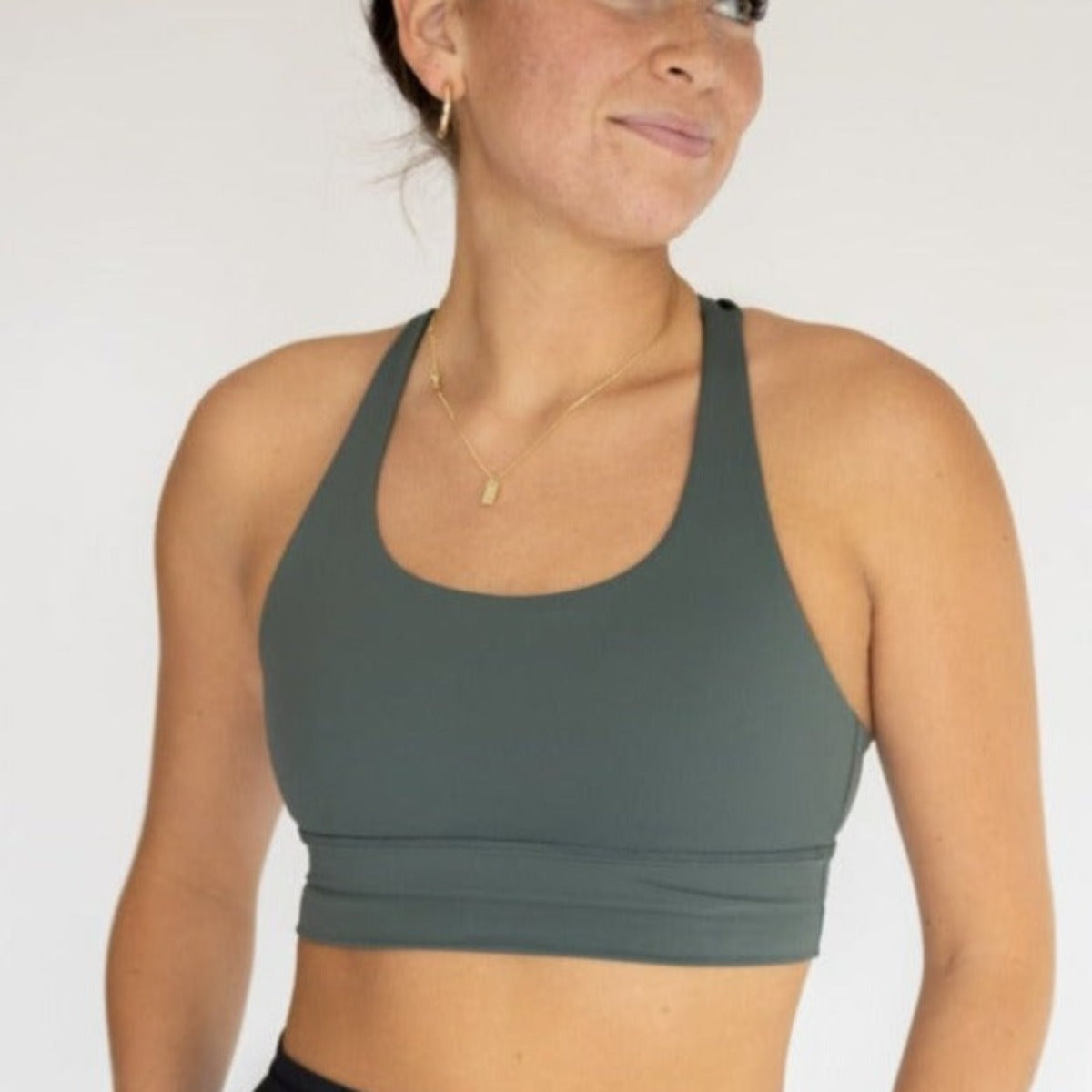 A woman with light skin and brown hair tied back wears the DYM Athletics Zone Bra | A-D+ cup | High Support, paired with gold hoop earrings and a necklace. She smiles slightly, looking to the side against a plain light background. Only her upper body is visible.