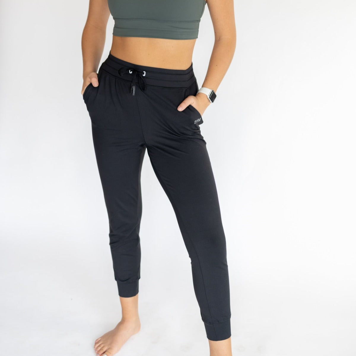 A woman stands barefoot against a white background, wearing DYM Athletics’ Performance Pro Lightweight Active Jogger with pockets and drawstring. She pairs it with a dark green crop top and smartwatch, hands in her pockets, face out of frame.