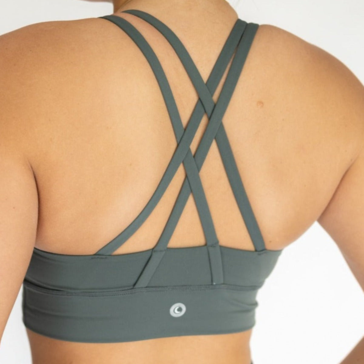 A woman is seen from behind wearing DYM Athletics Zone Bra | A-D+ cup | High Support in sage green, featuring multiple thin crisscross straps and a fitted, moisture-wicking band set against a plain light background.