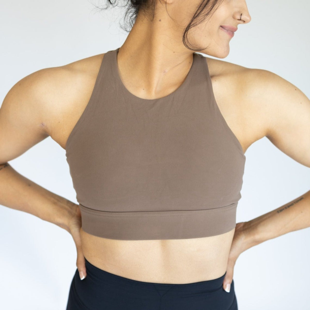 A dark-haired woman with her hair tied up smiles slightly, looking to the side. She wears the DYM Athletics AIRE Ultra-Soft Dynamic Adjustable Bra (high support, A-D+ cup) in brown and black leggings against a plain light background.