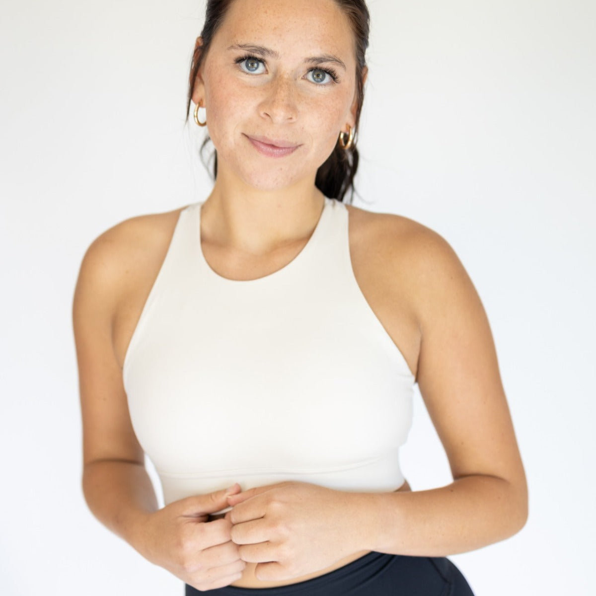 A woman with light skin, blue eyes, and dark brown hair stands against a white background wearing DYM Athletics' AIRE Ultra-Soft Dynamic Adjustable Bra (High Support, Cream) with a high neck and clasp, dark pants, gold hoops, and a confident smile.