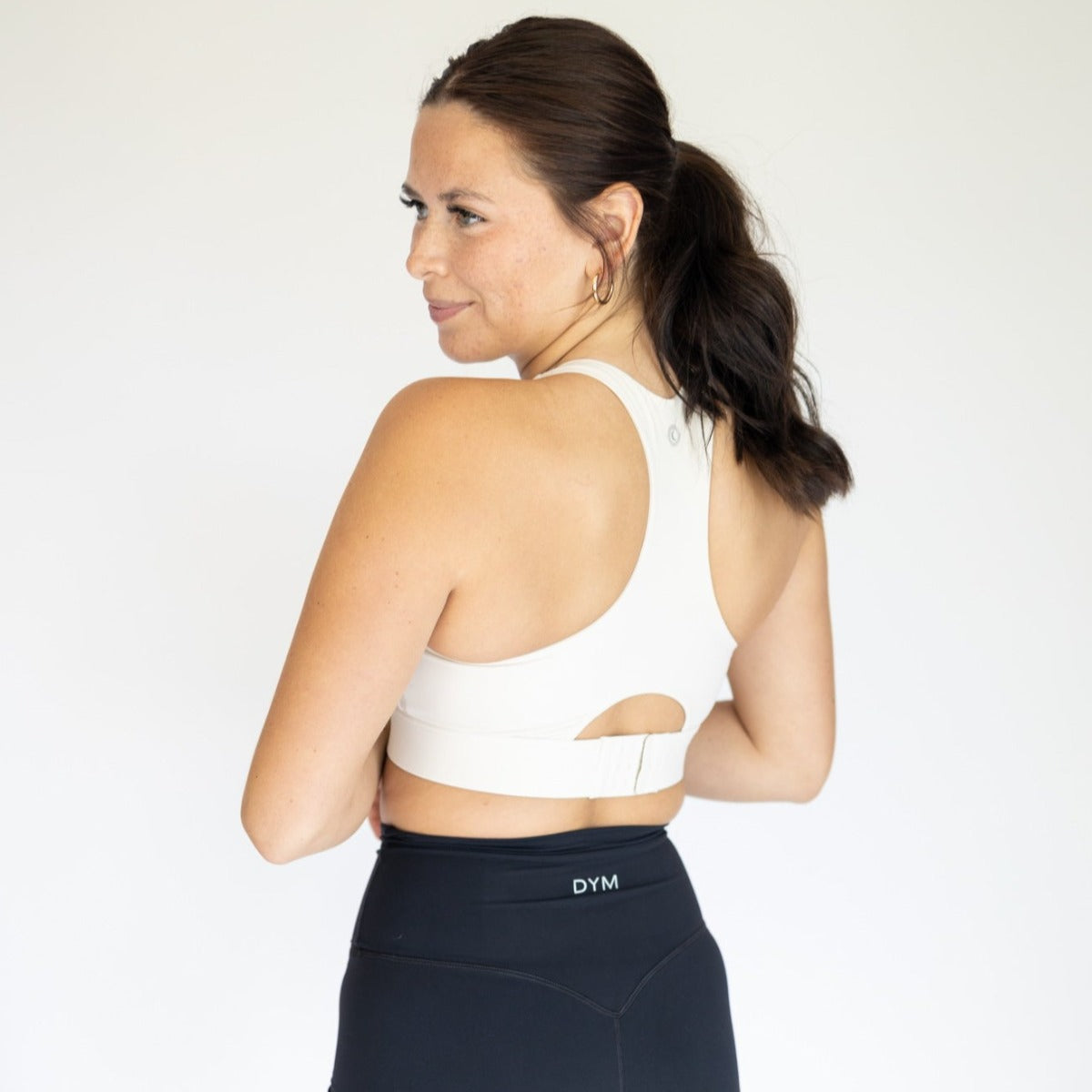 A woman with dark hair in a ponytail wears the DYM Athletics AIRE Ultra-Soft Dynamic Adjustable Bra (High Support) and high-waisted black leggings with a DYM logo, standing sideways and smiling slightly against a plain light background.