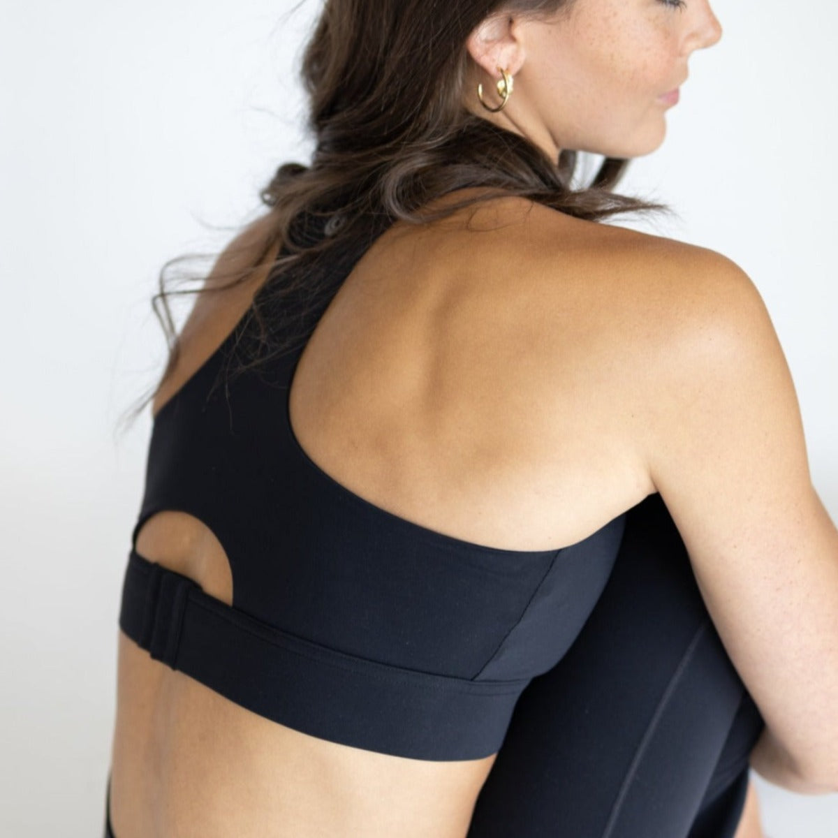 A woman with wavy brown hair and gold hoops sits turned from the camera, wearing the DYM Athletics AIRE Ultra-Soft Dynamic Adjustable Bra (high support, A-D+ cup) in black and matching leggings, against a white background.