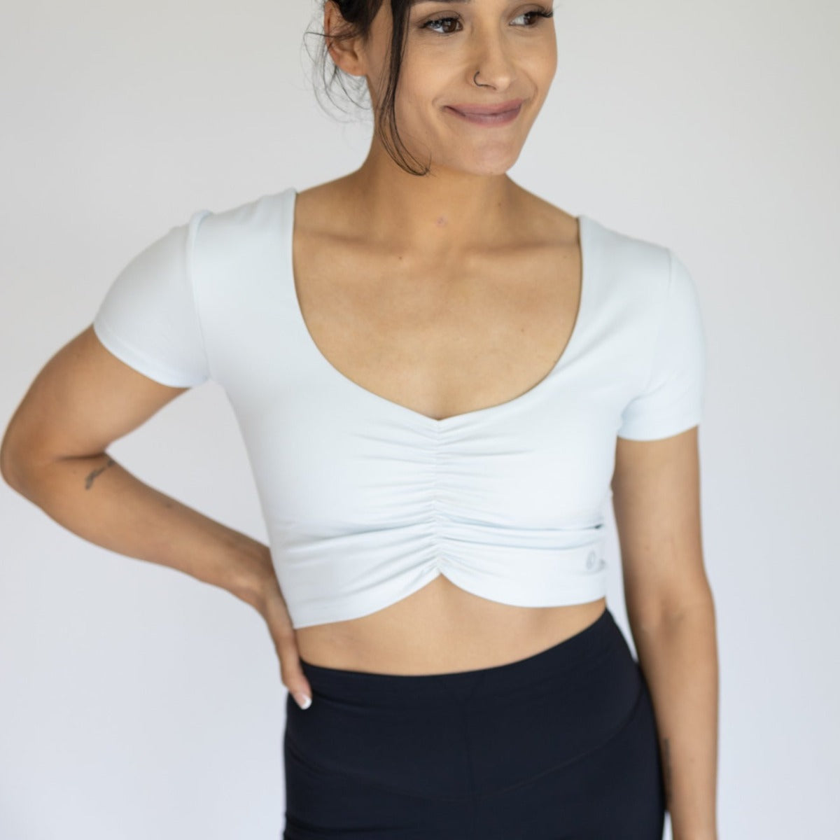 A woman with medium skin tone wears the DYM Athletics Bliss Two-Way Short Sleeve Top in a light color and high-waisted black leggings. She smiles softly, looking slightly to the side with one hand on her hip against a plain background.