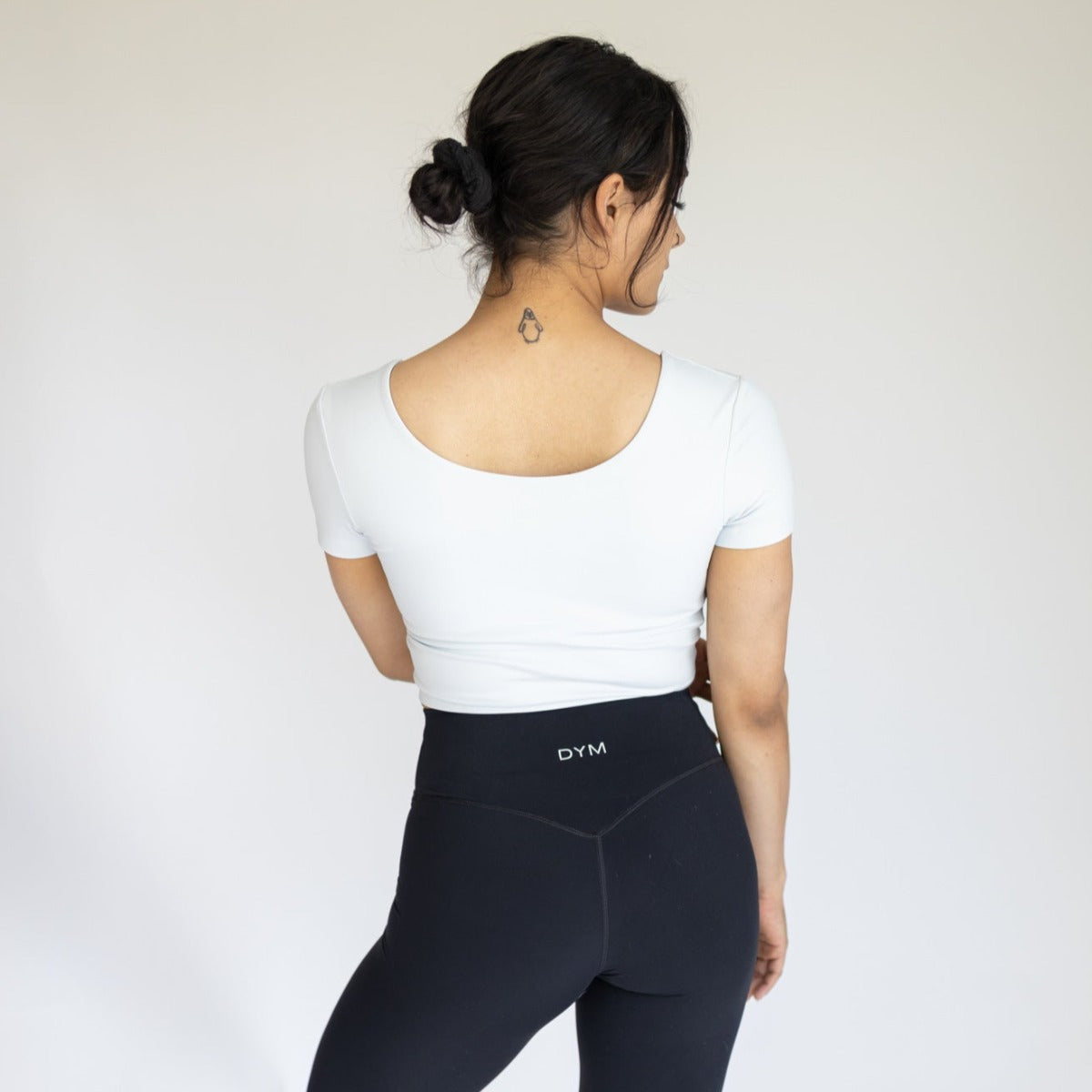 A woman with dark hair in a low bun faces away, wearing the DYM Athletics Bliss Two-Way Short Sleeve Top and high-waisted black leggings with a “DYM” waistband. A neck tattoo is visible against a plain, light background.