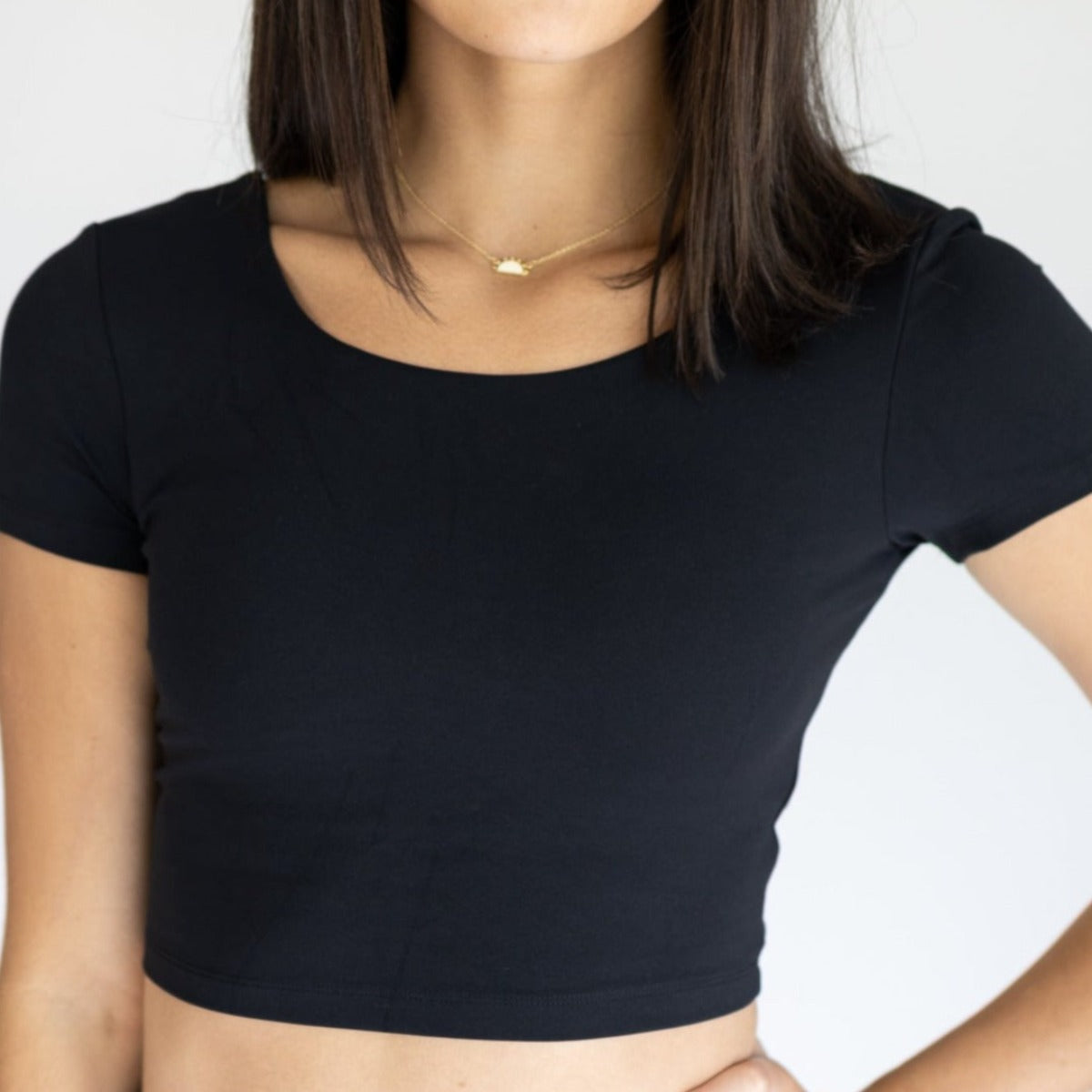 A woman models the DYM Athletics Bliss Two-Way Short Sleeve Top in black with a gold pendant necklace, standing against a white background. Her straight, shoulder-length dark hair and face are partially out of frame.