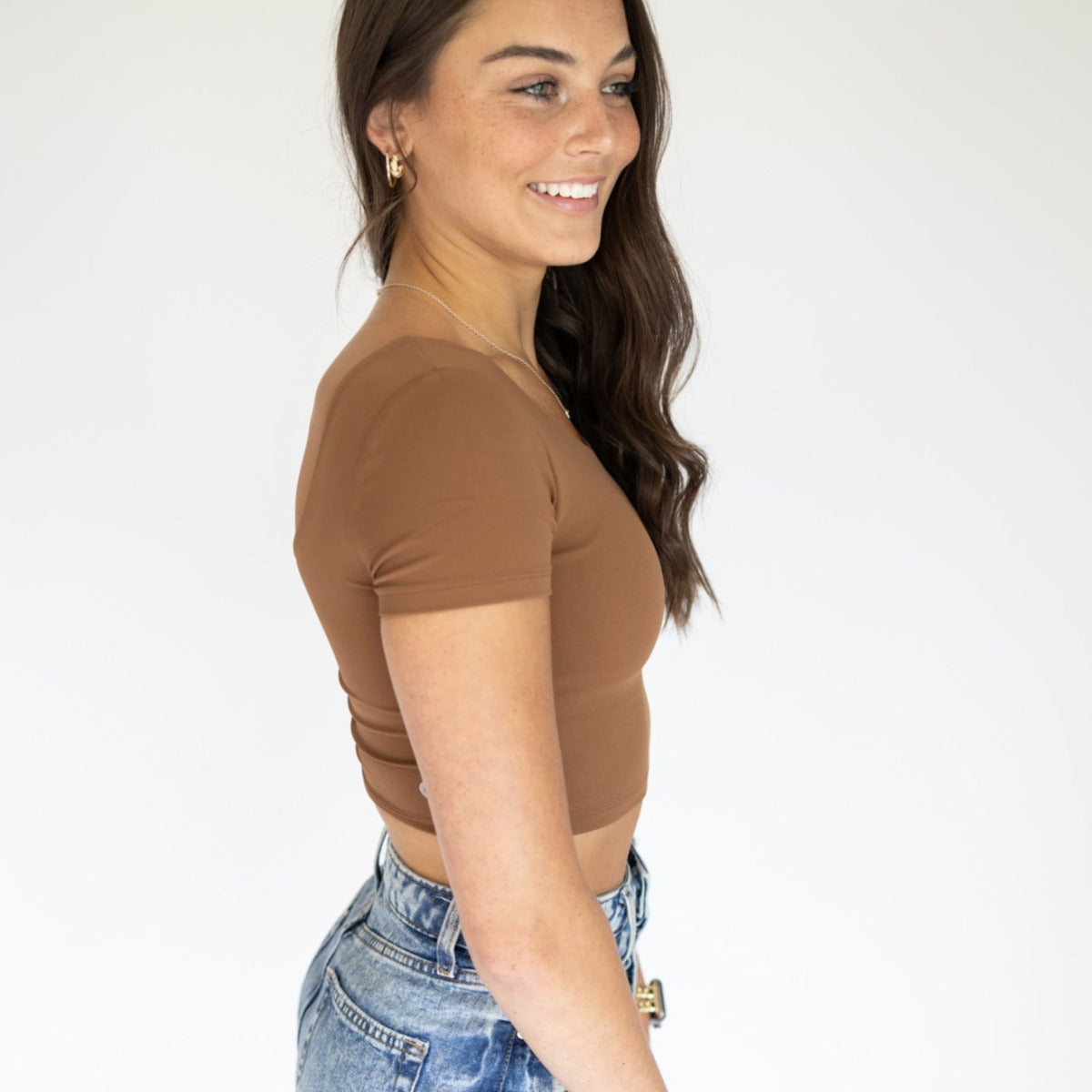 A woman with long brown hair stands in profile against a white background, smiling. She wears the DYM Athletics Bliss Two-Way Short Sleeve Top (SALE) with high-waisted blue jeans, a belt, gold hoop earrings, and a necklace.