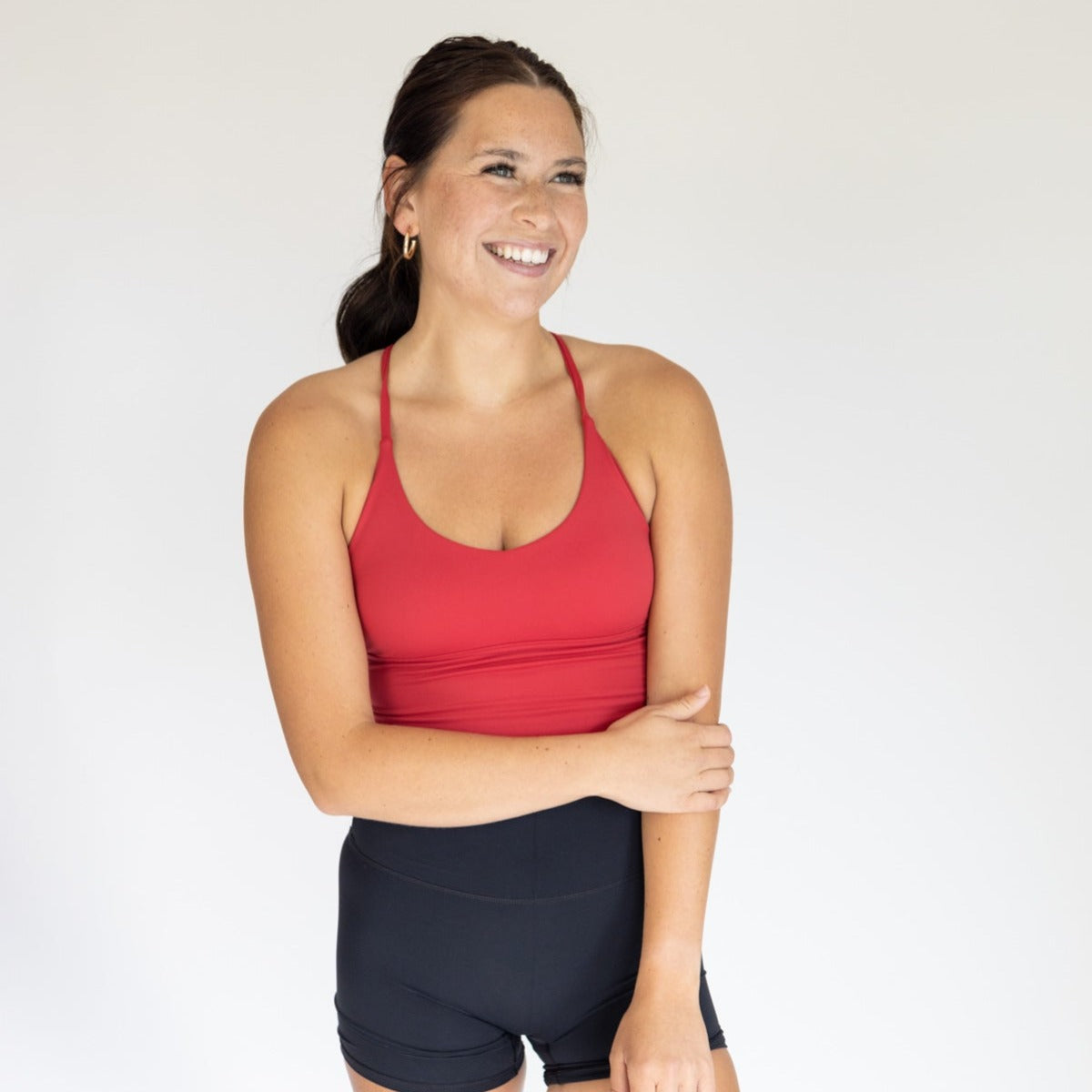 A woman with long brown hair in a ponytail smiles against a light background, wearing the DYM Athletics Flex & Flow Racerback Tank in red with extended tummy coverage and black high-waisted shorts, arms comfortably resting over her stomach.