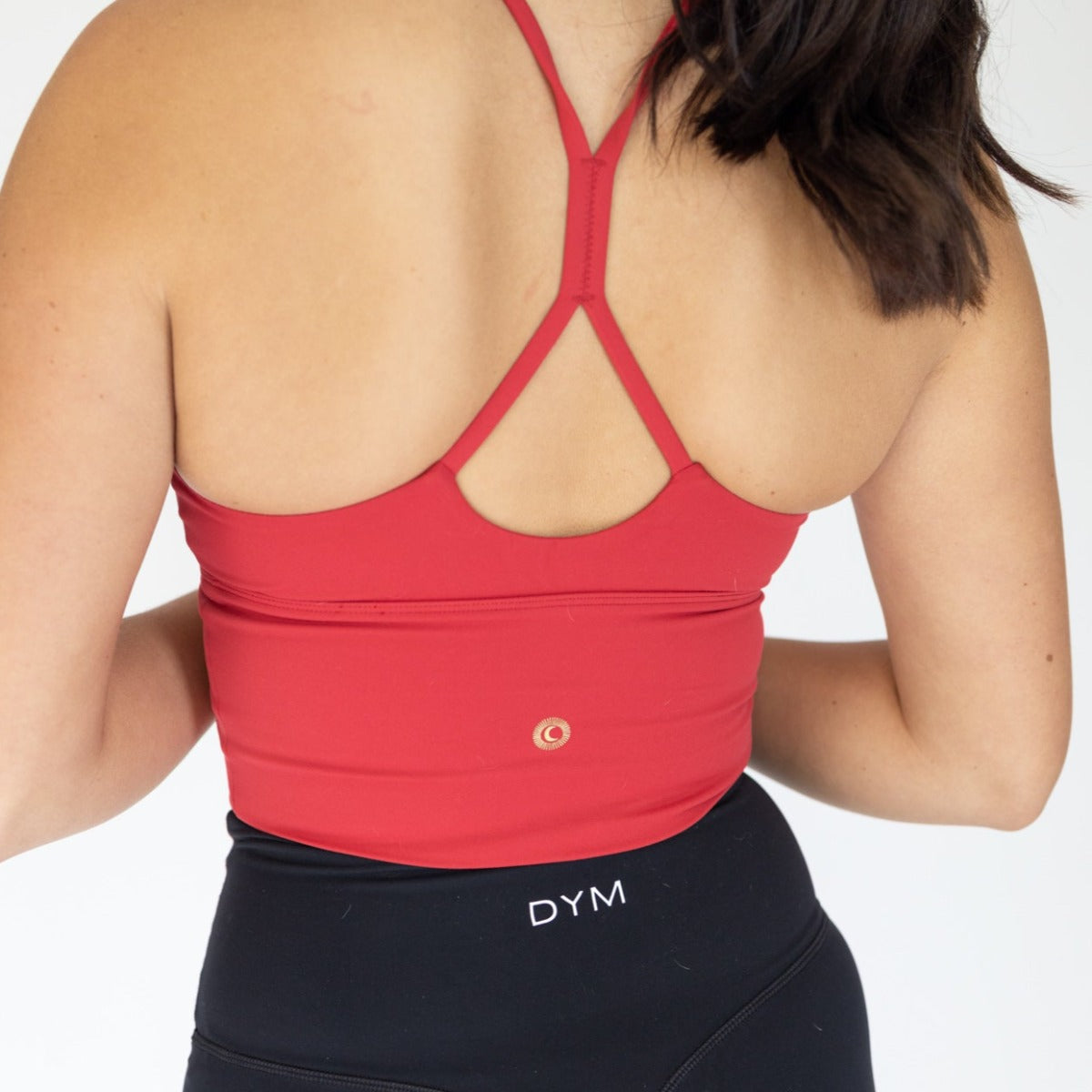 A person with long dark hair is shown from the back wearing a red Flex & Flow Racerback Tank and high-waisted black leggings with "DYM" on the waistband. Both items are from DYM Athletics, set against a plain white background.