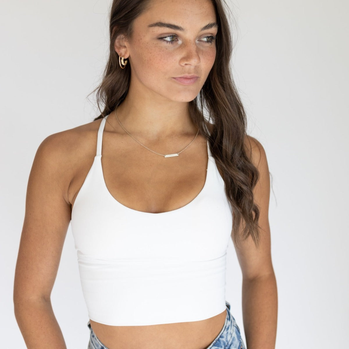 A young woman with long brown hair and light skin wears the DYM Athletics Flex & Flow Racerback Tank, featuring extended tummy coverage, paired with blue jeans. She stands against a light background, accessorized with a necklace and gold hoop earrings.