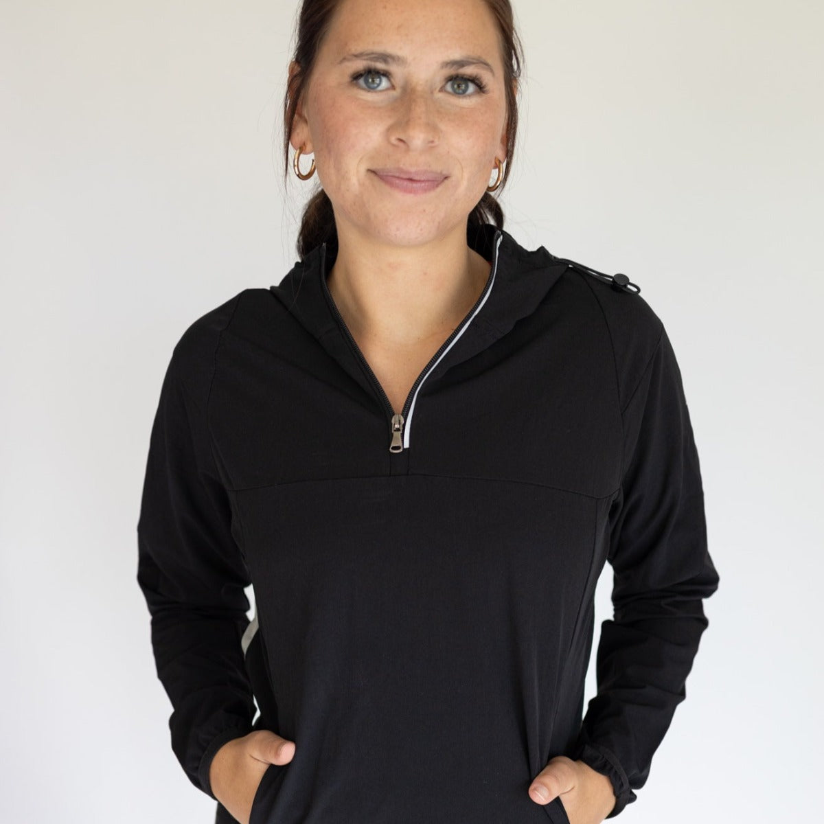 A woman with light skin and brown hair tied back smiles gently at the camera, wearing the DYM Athletics Women's Hiking Jacket (SALE) in black. She has gold hoop earrings, hands in her pockets, and the background is plain and light-colored.