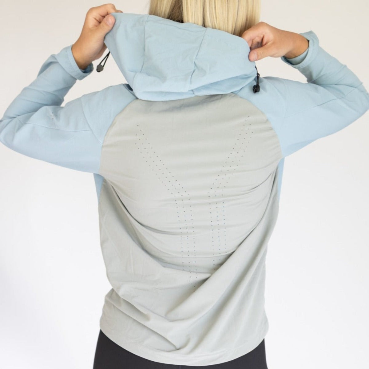 A woman with blonde hair faces away, wearing the DYM Athletics Women's Hiking Jacket in light blue and gray. The hood is up, held by both hands, showing perforated lines on the back. Background is plain white. Now on SALE!.
