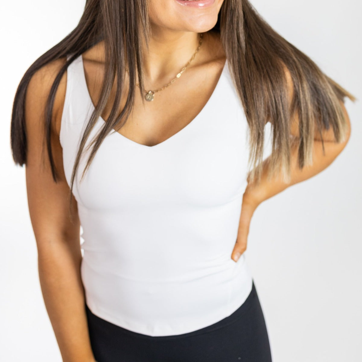 A woman with straight brown hair wears the DYM Athletics Bliss"Full" V-Neck Tank with Scoop Back (A-D Cup, SALE) in buttery soft fabric, paired with black pants. She smiles, hand on hip, against a white background; gold necklace visible. Face partly out of frame.