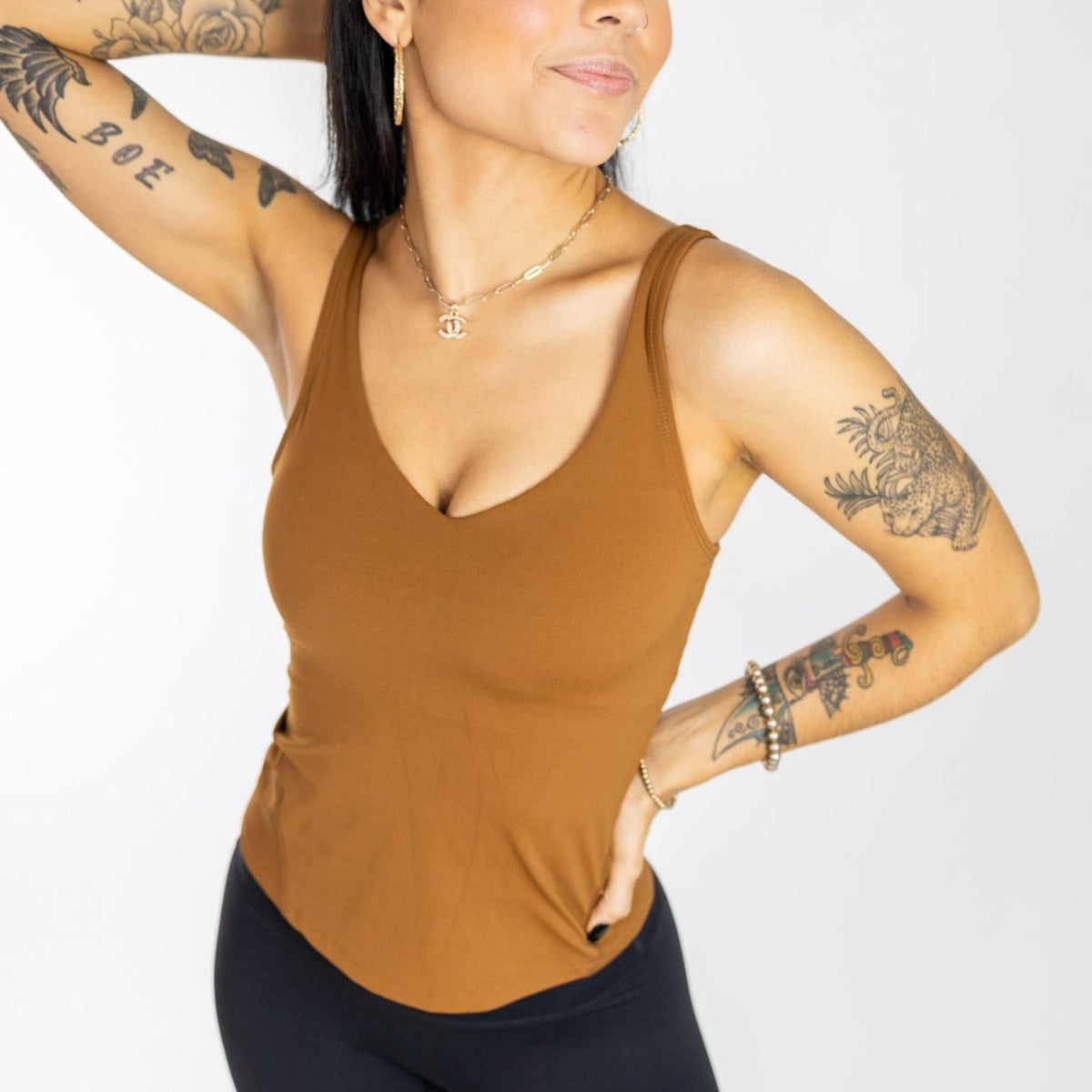 A woman with long dark hair and visible tattoos models the Bliss"Full" V-Neck Tank with Scoop Back by DYM Athletics (A-D Cup, SALE), paired with black pants, gold jewelry, and stands confidently against a plain white background.
