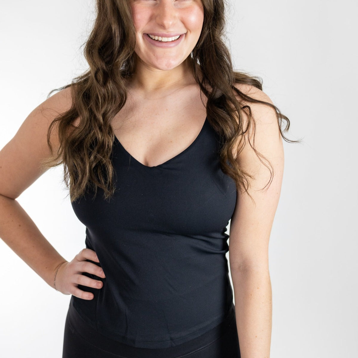 A woman with long wavy brown hair smiles in a DYM Athletics Bliss"Full" V-Neck Tank with Scoop Back (A-D Cup), paired with black pants. She stands against a white background, hand on hip. The buttery soft tank features a built-in shelf bra for support.