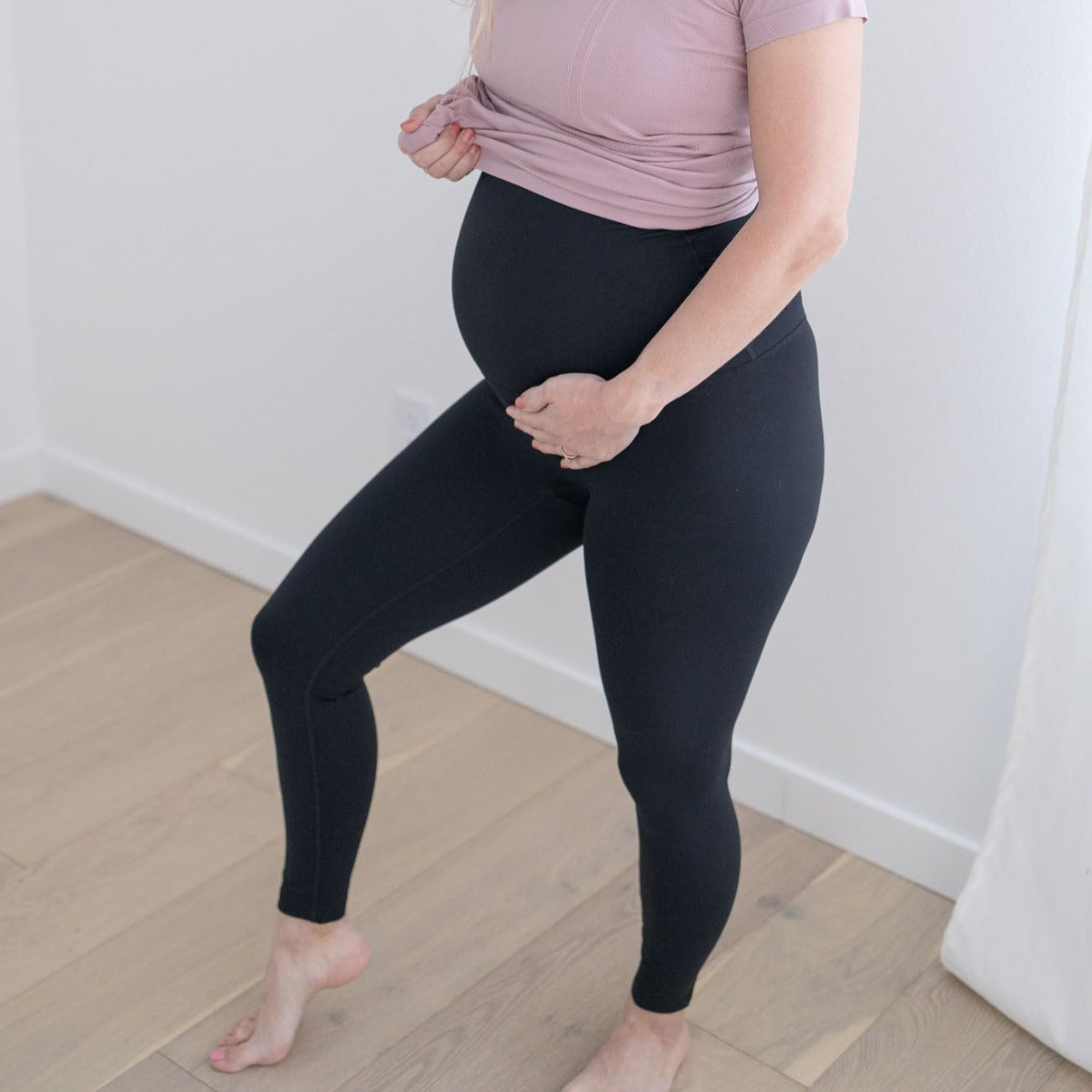 A pregnant woman stands barefoot on a light wooden floor, wearing DYM Athletics' LOVE THE BUMP Maternity Belly Support Legging and a mauve top, gently lifting her shirt to show her baby bump against a backdrop of white walls and curtains.