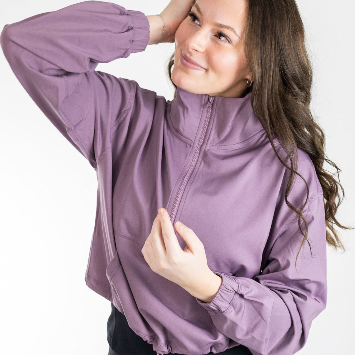A smiling woman with long brown hair models the DYM Athletics Ascend 1/4 Zip Jacket in mauve. She stands against a white background, raising one hand to her head and holding the jacket’s hem while looking slightly upwards.