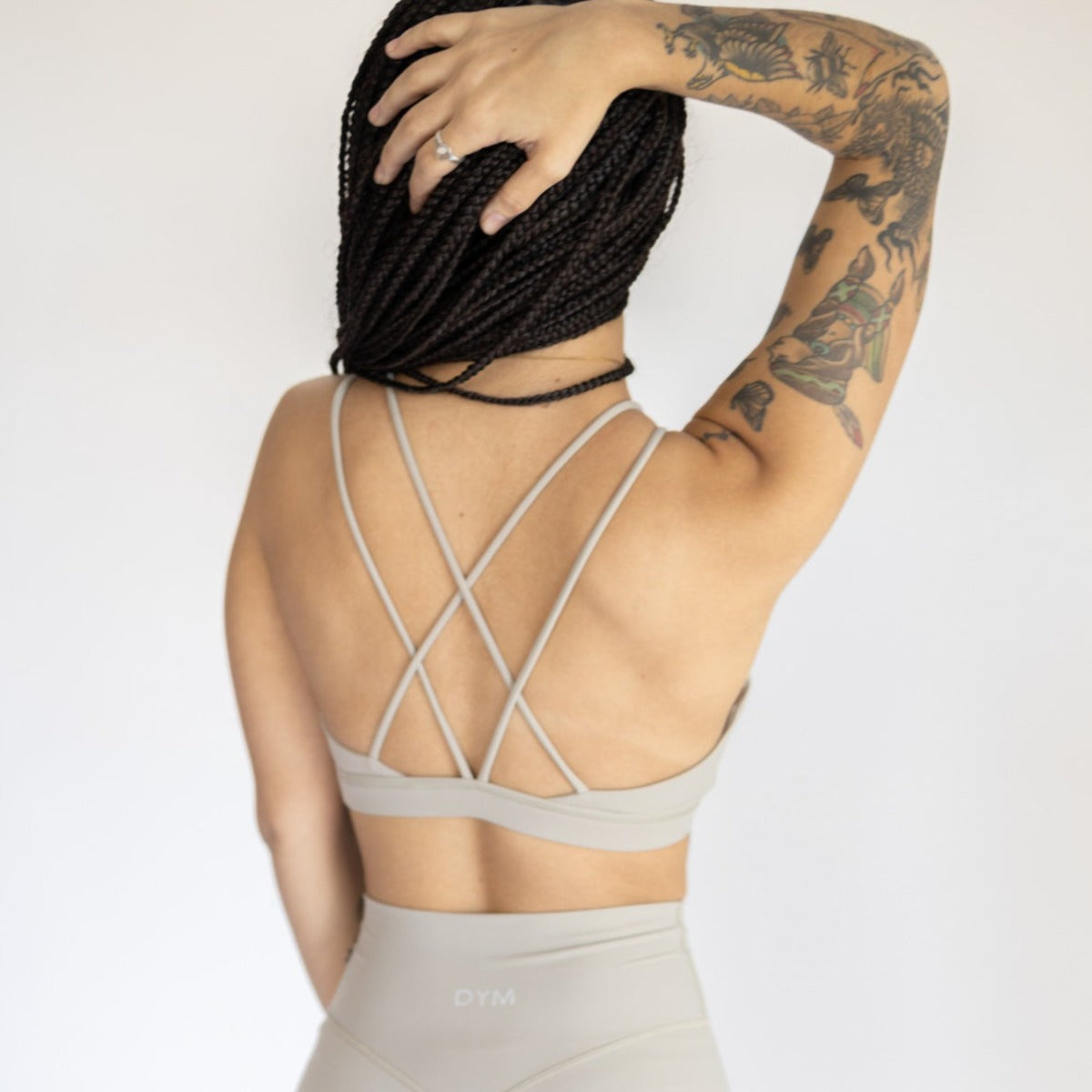 A person with braided hair faces away, wearing the DYM Athletics AIRE Ultra-Soft Cross Back Sports Bra in light beige and matching leggings. Their left arm is raised, displaying upper arm tattoos against a plain white background.