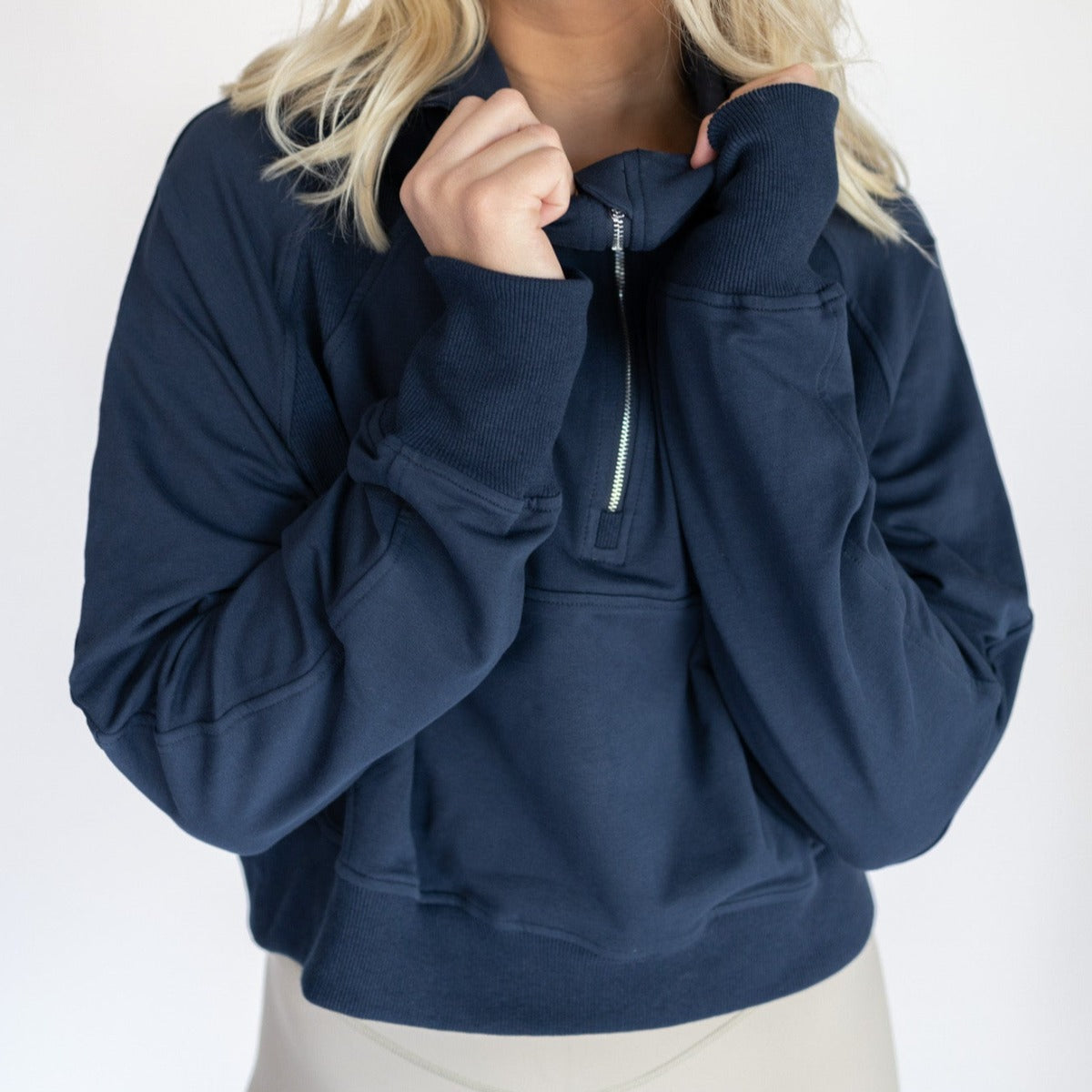 A person with long blond hair wears the DYM Athletics Lightweight 1/4 Zip Hoodie | SALE in navy blue, featuring thumbholes. They hold the collar near their face against a plain, light background. The image is cropped at mouth and waist.