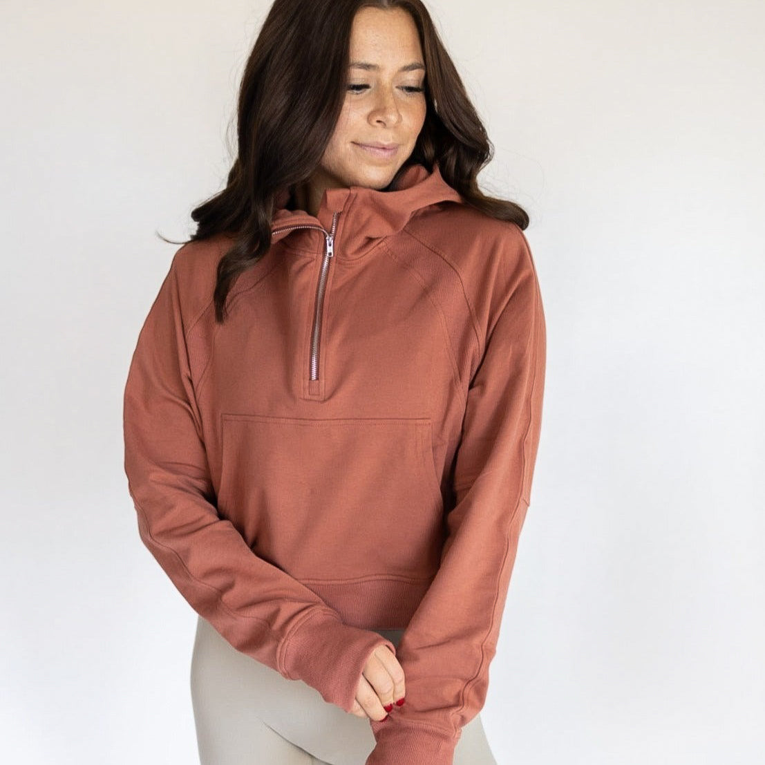 A woman with long brown hair wears the DYM Athletics Lightweight 1/4 Zip Hoodie in rust and light beige leggings, standing against a plain background, looking down to her left with a relaxed expression and her hands gently clasped.