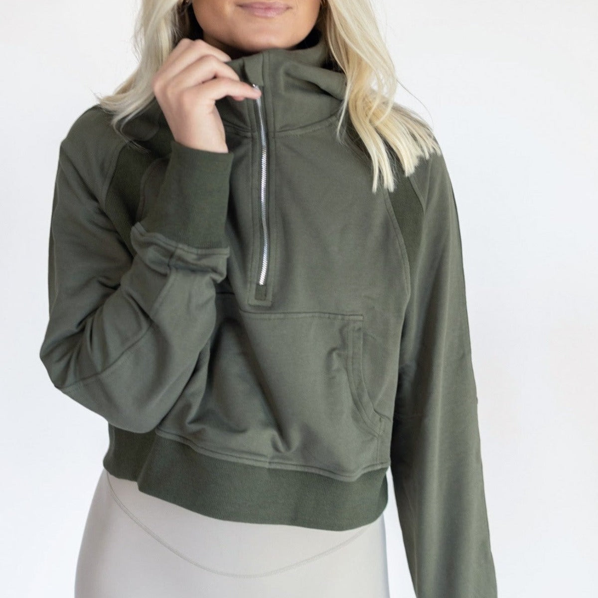 A person with long blonde hair models the DYM Athletics Lightweight 1/4 Zip Hoodie in olive green. Featuring a high collar and cropped fit, the stylish, long-sleeve hoodie stands out against a light background; only their torso and lower face are shown.