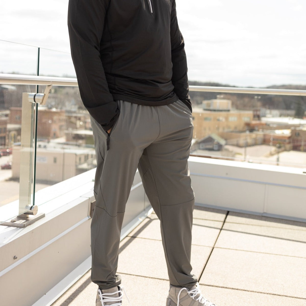 A person stands on a rooftop with hands in pockets, wearing a black long-sleeve top, DYM Athletics Fly Lightweight Active Jogger in gray, and light gray sneakers. A glass railing frames the cityscape and clear sky behind them.