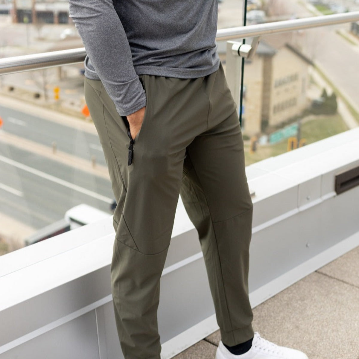 A person stands on a balcony wearing DYM Athletics’ Fly Lightweight Active Jogger in olive green, paired with a gray long-sleeve shirt and white sneakers. Their left hand rests in their pocket, city streets and buildings softly visible below.