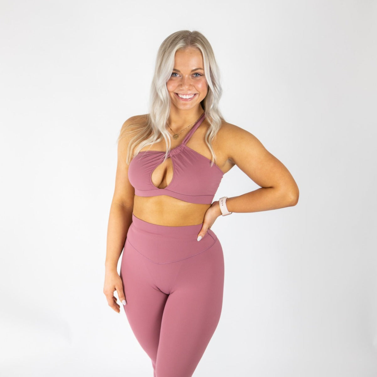 A woman with long, wavy platinum blonde hair smiles against a white backdrop. She wears mauve Muse Leggings and the DYM Athletics Siren One Shoulder Cutout Bra (A-C cup)—perfect for barre or stylish everyday wear. A white wristwatch accents her look.