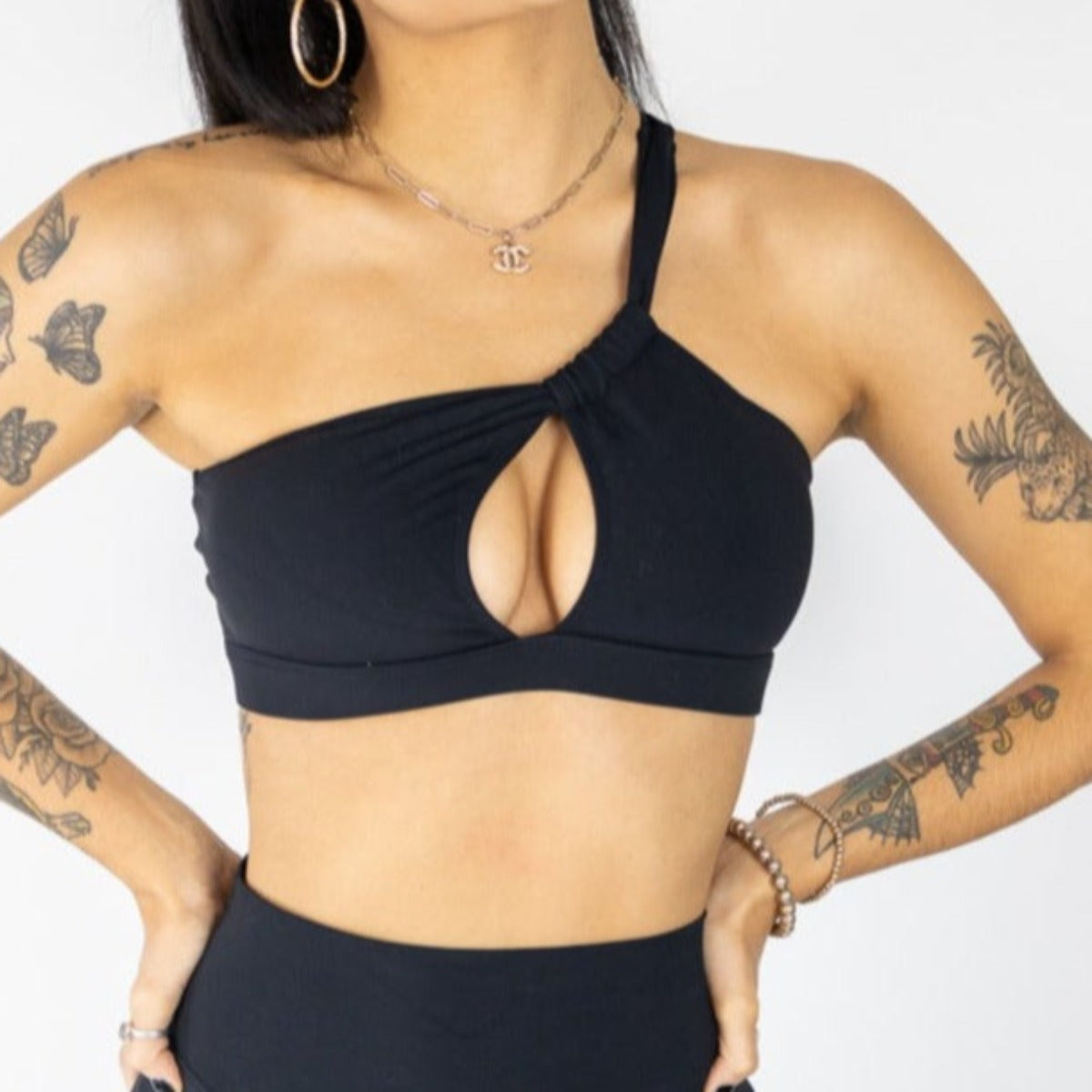 A woman wears DYM Athletics' The Siren One Shoulder Cutout Bra (A-C Cup) with Muse Leggings, accessorized with hoop earrings, layered necklaces, and bracelets—an effortlessly chic everyday look.