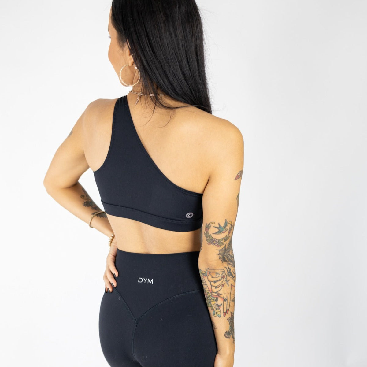 A woman with long dark hair and tattooed arms wears the DYM Athletics Siren One Shoulder Cutout Bra (A-C Cup, SALE) and Muse Leggings, perfect for Barre or daily wear, with “DYM” on the waistband, against a white background.