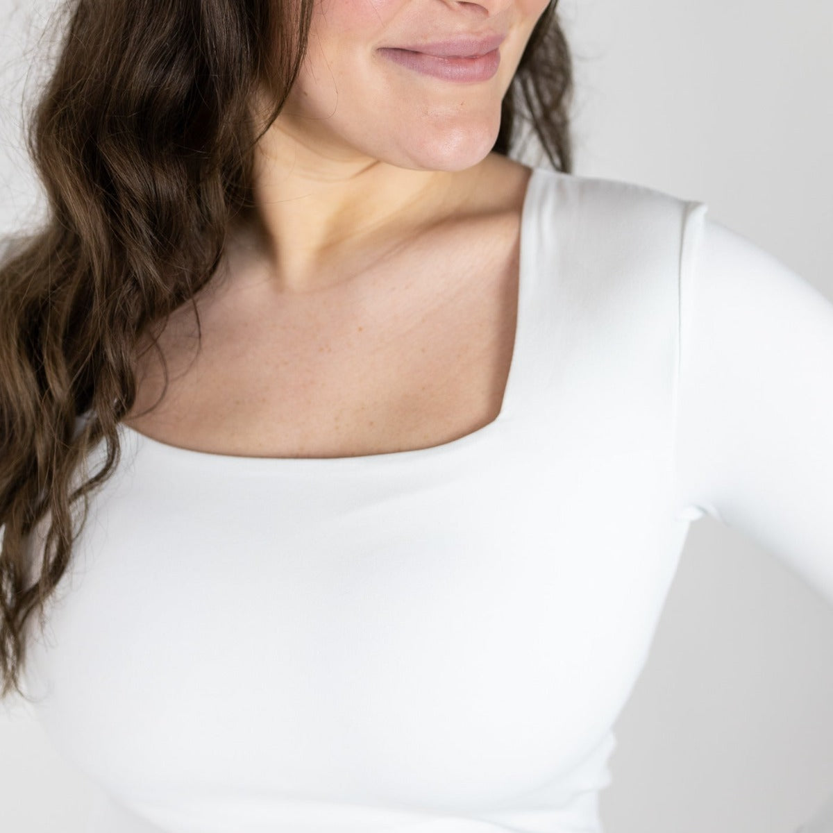 A close-up shows a woman with long brown wavy hair, smiling softly in the DYM Athletics Bliss Square Neck Long Sleeve Top—a fitted, breathable top with a square neckline. The plain, light background highlights her mouth, chin, neck, and upper torso.