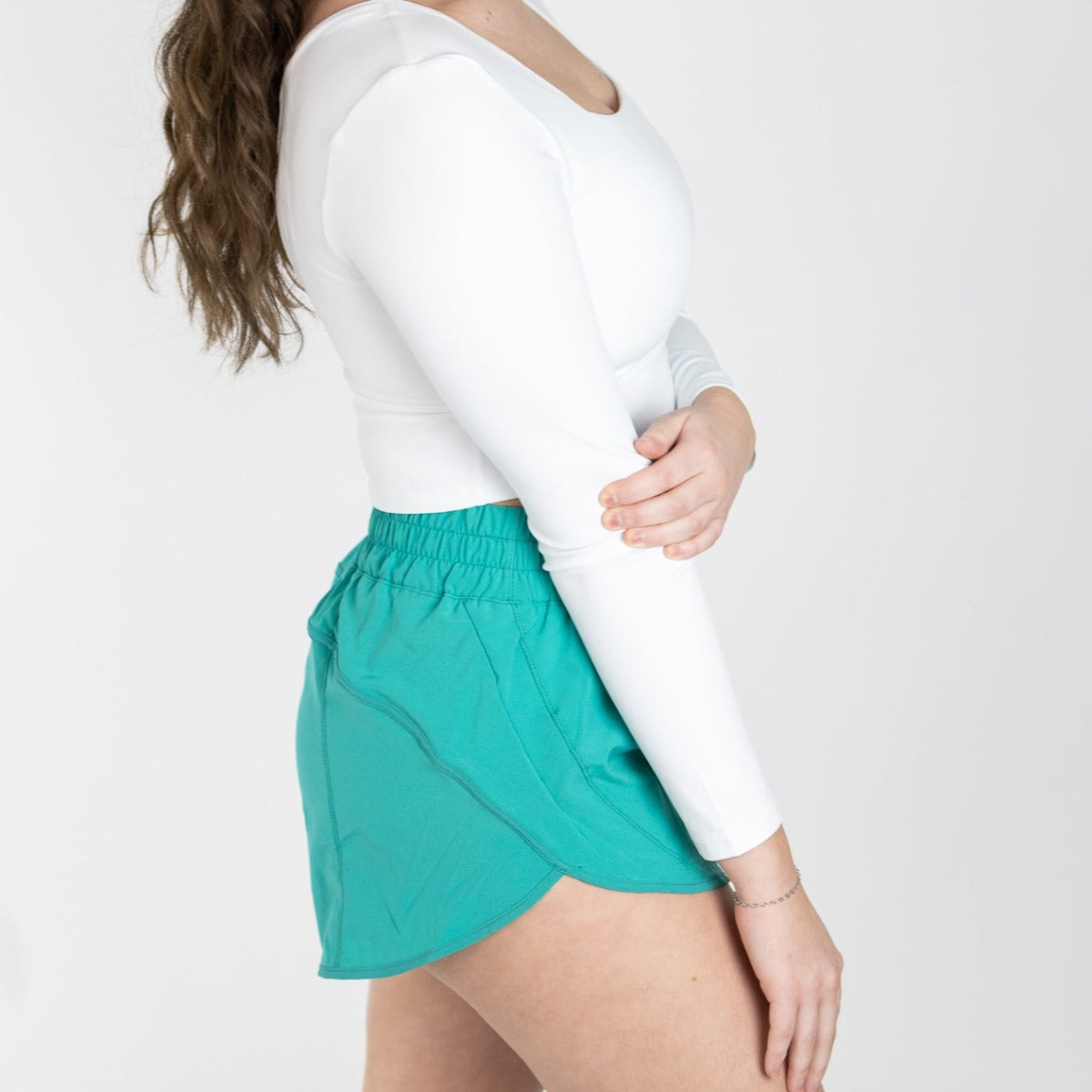 A woman stands sideways against a white background, wearing a fitted long-sleeve white top and DYM Athletics’ Sprint High-Rise Lined Short | 3" in teal. Her wavy brown hair falls over her shoulder, with her right arm bent and face not visible.