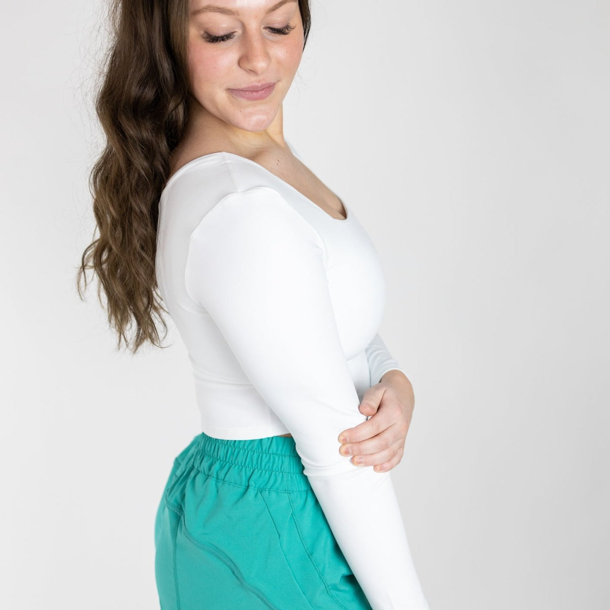 A woman with long wavy brown hair wears the DYM Athletics Bliss Square Neck Long Sleeve Top (SALE) with teal high-waisted pants, standing against a plain light background, gently holding her opposite elbow and smiling slightly.