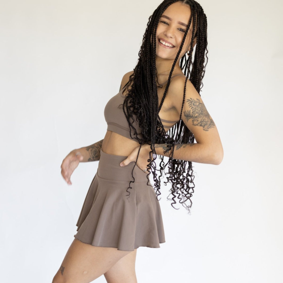A smiling woman with long, curly braids and visible tattoos models the DYM Athletics AIRE Ultra-Soft Tennis Skirt w/ Pockets on sale, paired with a matching taupe crop top, playfully posed against a plain light background.