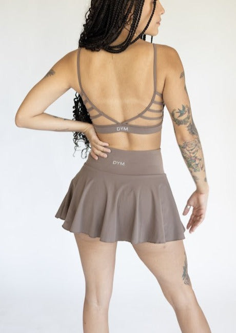 A woman with long braids and tattoos stands with her back to the camera, wearing a taupe DYM Athletics sports bra and the AIRE Ultra-Soft Tennis Skirt w/ Pockets, both featuring “DYM” logos. The plain background is light-colored.