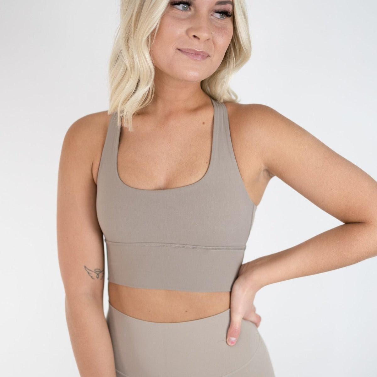 A woman with shoulder-length blonde hair models the DYM Athletics Vibe Bra with Detailed Back (medium-high support, A-D cup) in taupe, paired with matching high-waisted leggings. She stands against a white background, smiling softly, her forearm tattoo visible.