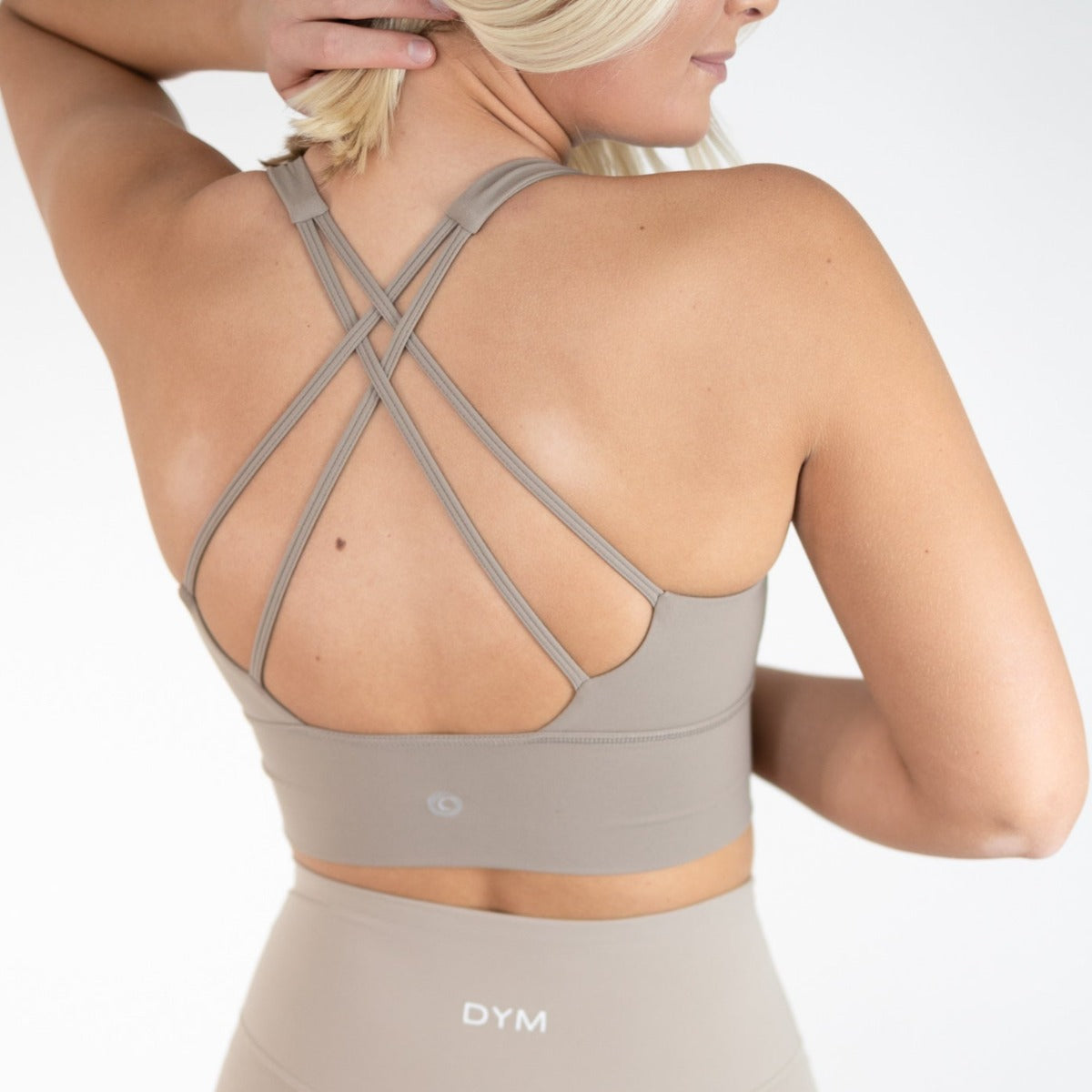 A blonde woman wears the DYM Athletics Vibe Bra with detailed back, a taupe, medium-high support sports bra with thin criss-cross straps and matching high-waisted leggings bearing “DYM” on the waistband, against a plain white background.
