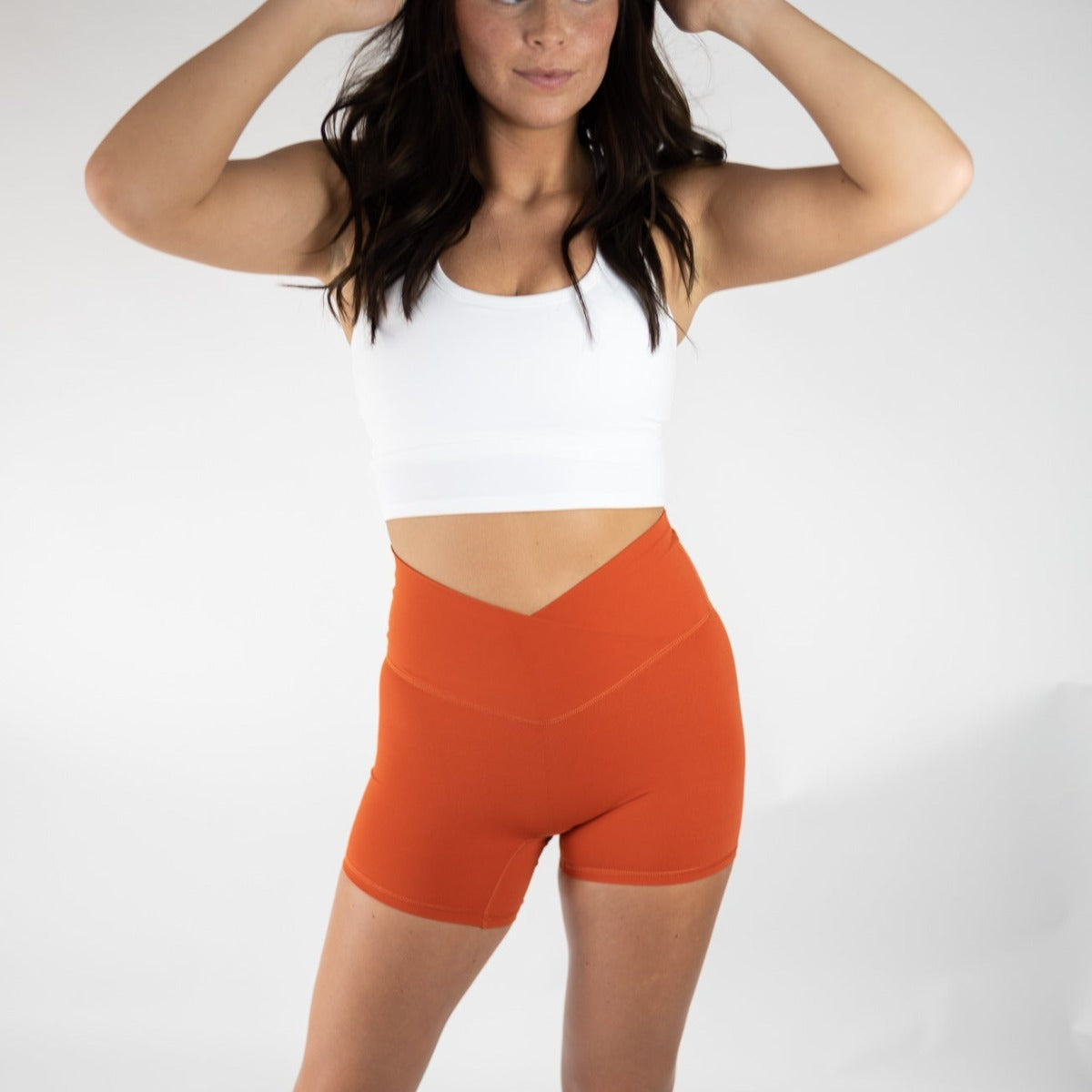 A woman in a white sports bra pairs it with DYM Athletics' Flex & Flow V-Waist Short | 4" | SALE in burnt orange, posing against a plain white background with her hands raised and long dark hair framing her partly hidden face.