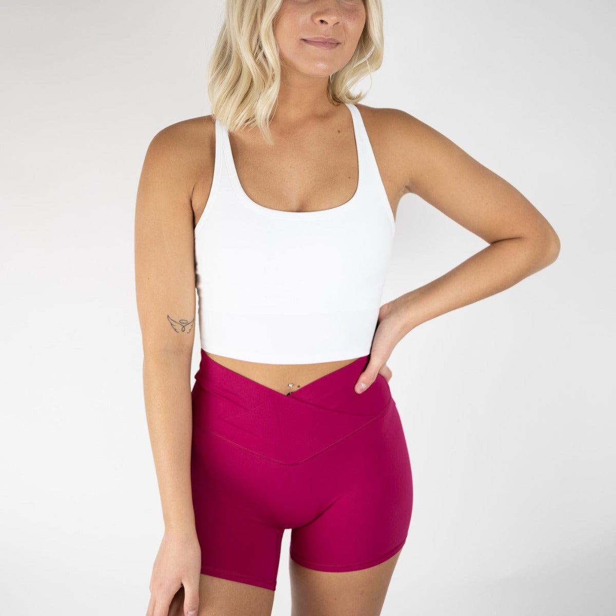 A blonde woman models the DYM Athletics Flex & Flow V-Waist Short | 4" (SALE) and a white crop tank top, standing against a plain white background with one arm on her hip, revealing a small tattoo on her left arm.