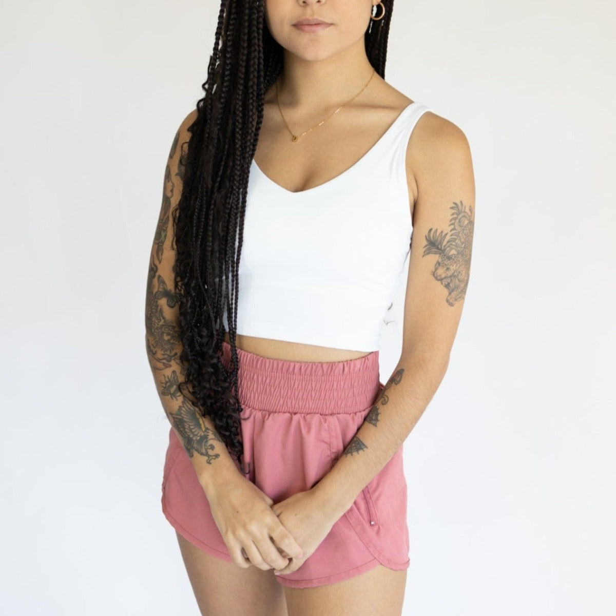 A person with long braids and arm tattoos stands against a plain background, wearing the DYM Athletics Pace High-Waisted Lined Short | 3" (SALE) in pink, paired with a white crop top, hoop earrings, and a delicate necklace.