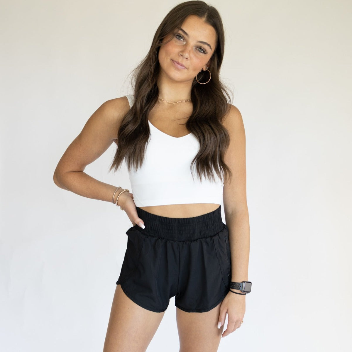 A young woman with long wavy brown hair poses against a plain backdrop, smiling softly. She wears a white crop top, hoop earrings, bracelets, a black smartwatch, and the Pace High-Waisted Lined Short | 3” by DYM Athletics—ideal for running.